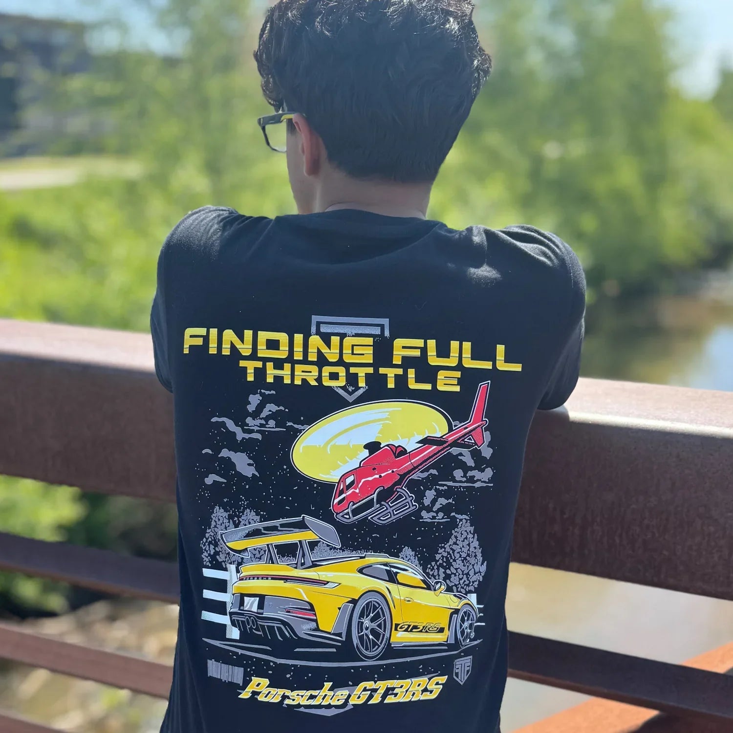 Person wearing a black t-shirt with a graphic design of a car and text, standing outdoors.
