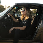 A woman with blonde wavy hair wearing a black t-shirt, black pants, and white sneakers sits in the driver's seat of a dark metallic Porsche. She is wearing sunglasses and looking to the side.