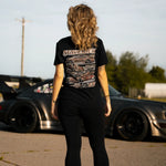 A woman stands with her back to the camera wearing a black graphic t-shirt and black leggings, standing in front of a dark gray sports car in an outdoor lot.
