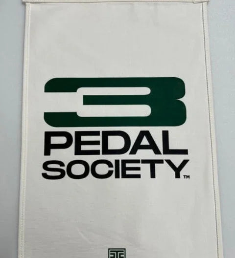 An off-white canvas flag featuring the dark green and black 'Pedal Society' logo, with metal grommets on the left side, shown against a neutral gray background.