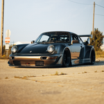 A dark metallic Porsche 911 with a widebody kit, lowered suspension, and a rear spoiler, parked on an outdoor concrete surface with a blurred background of sky and utility poles.