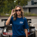 A woman wearing a blue short-sleeve t-shirt with a white 'SPEED SPORT' logo stands in front of a dark sports car outdoors.