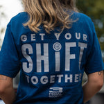 A person viewed from behind wearing a blue graphic T-shirt with large white text reading 'GET YOUR SHIFT TOGETHER' and a small 'PEDAL SOCIETY' logo near the bottom, set against a blurred outdoor background.