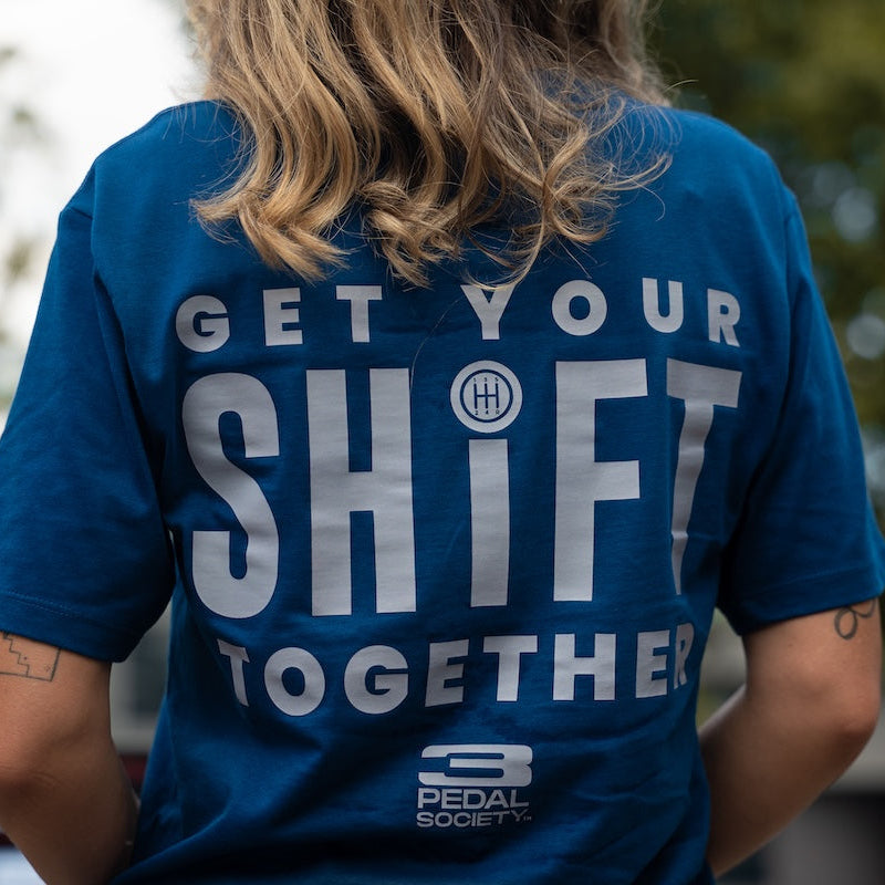 A person viewed from behind wearing a blue graphic T-shirt with large white text reading 'GET YOUR SHIFT TOGETHER' and a small 'PEDAL SOCIETY' logo near the bottom, set against a blurred outdoor background.