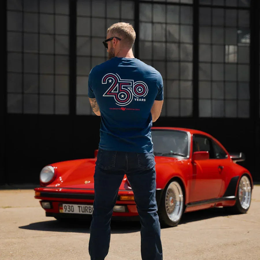 Man wearing a blue t-shirt with '250' logo standing next to a red sports car in front of a large window.
