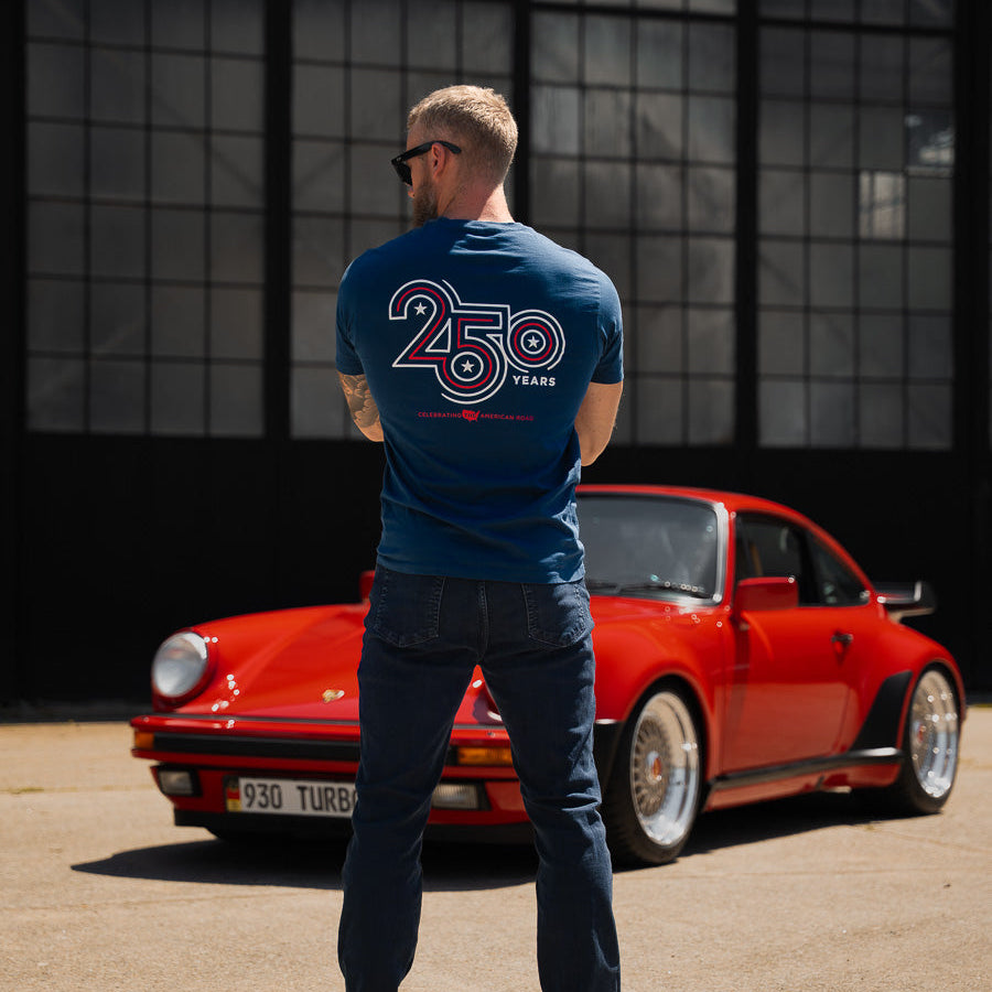 Man wearing a blue t-shirt with '250' logo standing next to a red sports car in front of a large window.