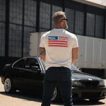 Man wearing a white t-shirt with a logo standing in front of a black car.