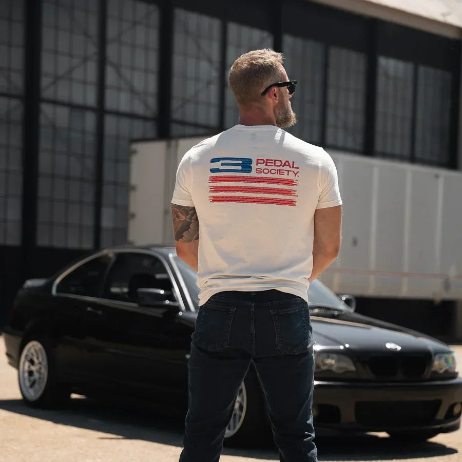 Man wearing a white t-shirt with a logo standing in front of a black car.
