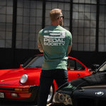 Man wearing a 'Pedal Society' t-shirt standing between two cars with a geometric wall in the background.