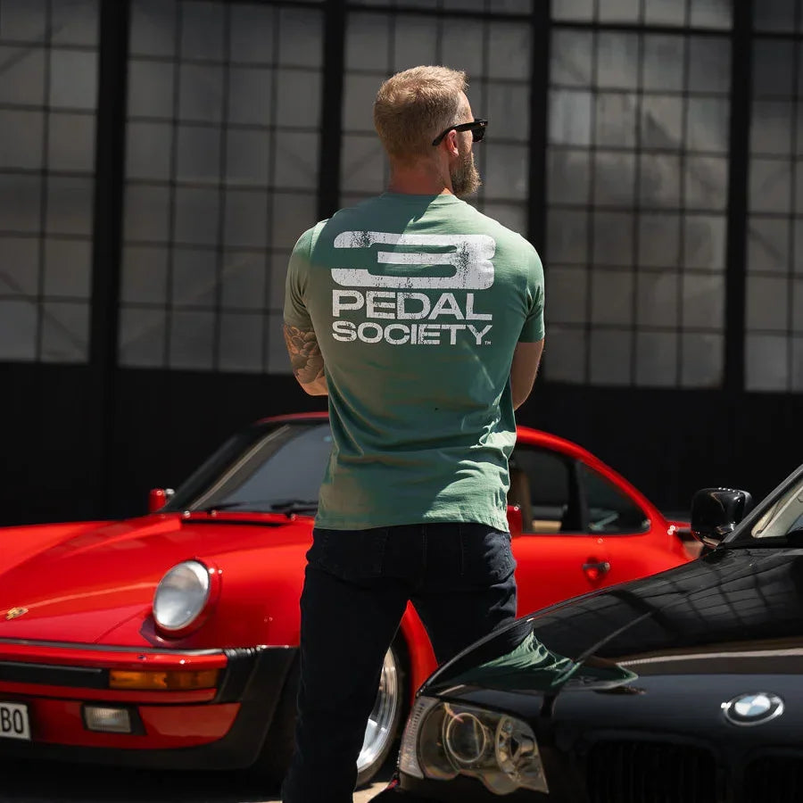 Man wearing a 'Pedal Society' t-shirt standing between two cars with a geometric wall in the background.