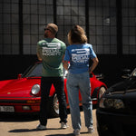 Two people wearing 'Pedal Society' shirts standing next to a red car in front of a grid-patterned wall.