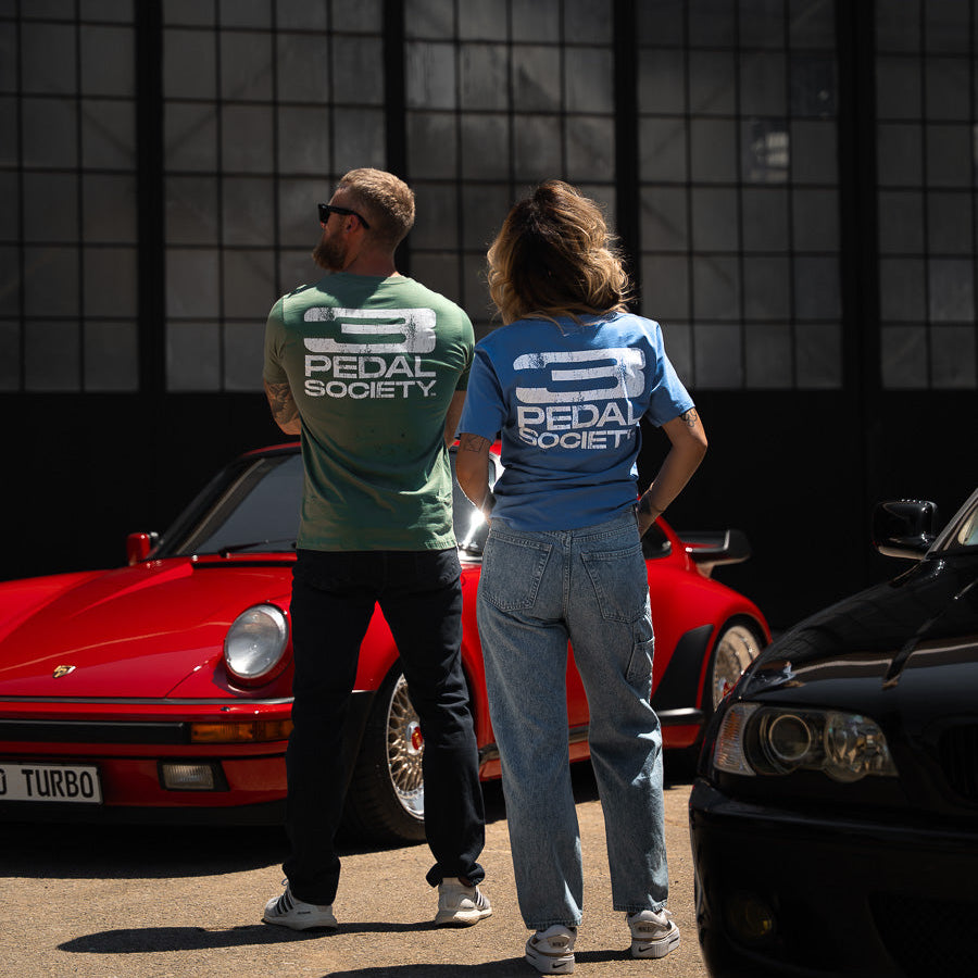 Two people wearing 'Pedal Society' shirts standing next to a red car in front of a grid-patterned wall.