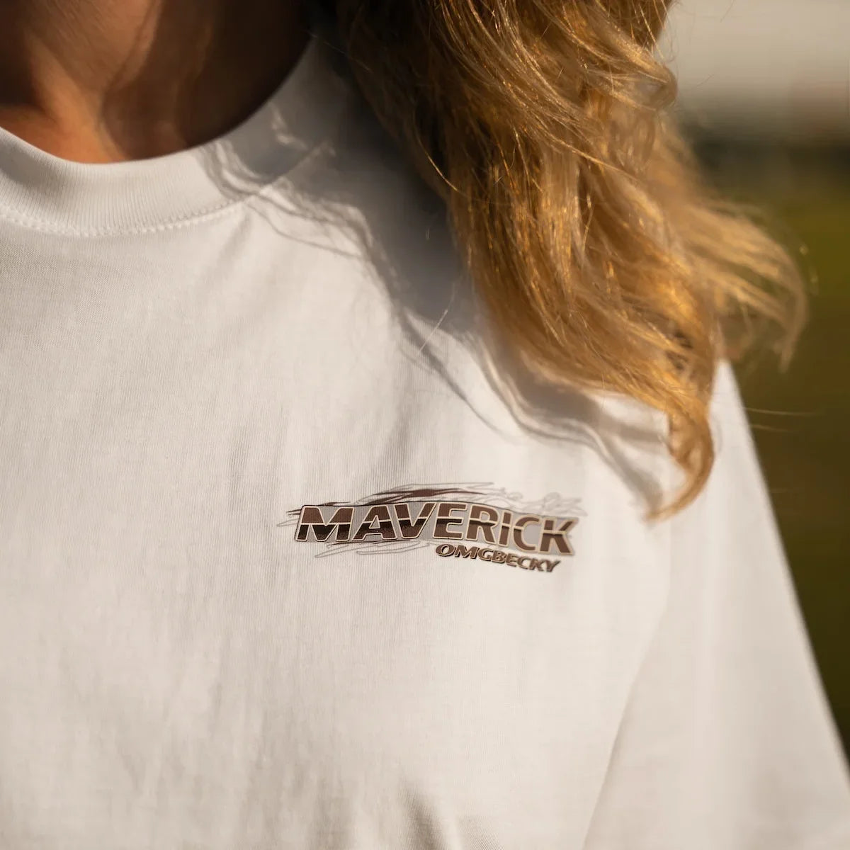 A close-up of a person wearing a white crewneck t-shirt with a brown and black 'MAVERICK GAWLICKY' logo printed on the left chest, set against a blurred outdoor background.