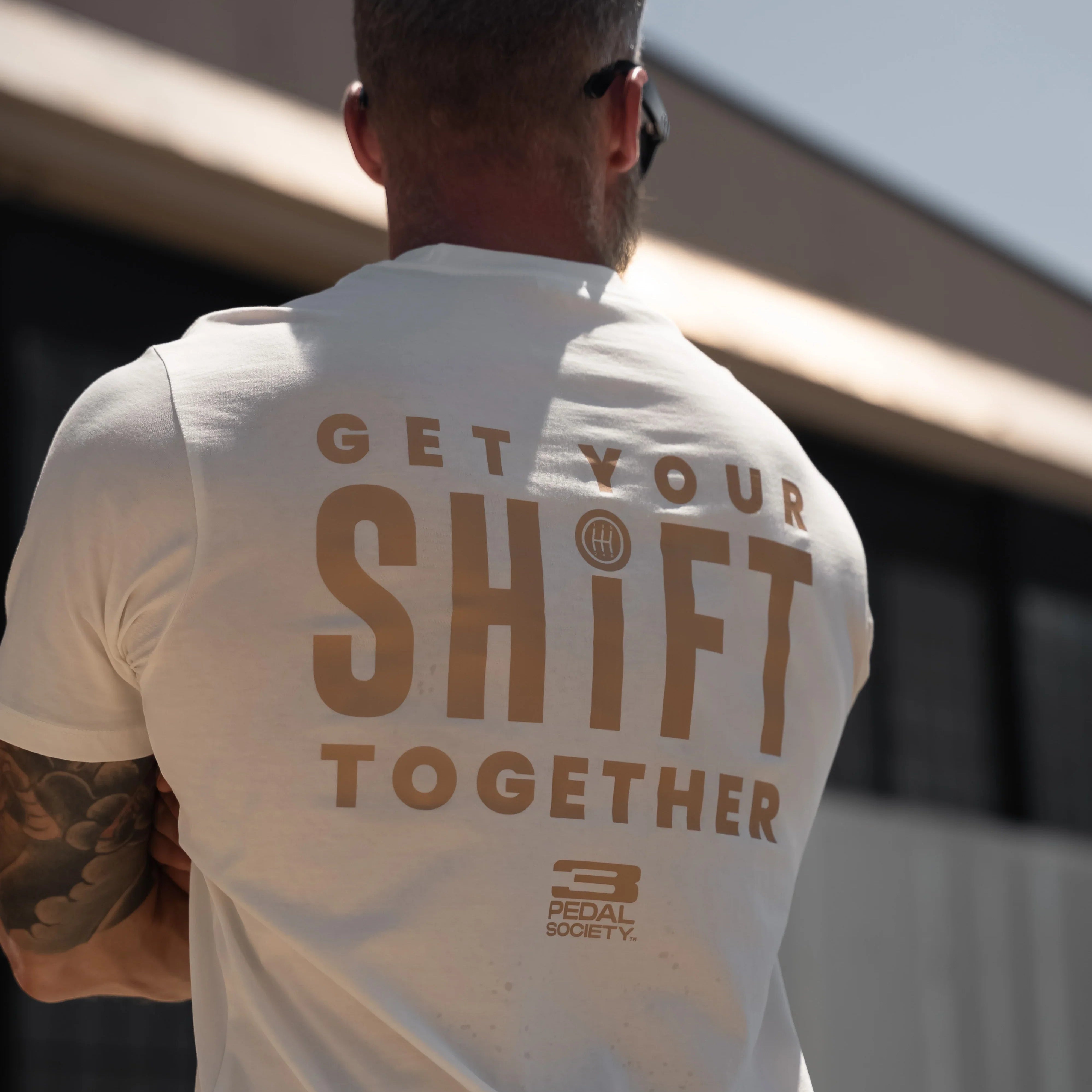 A man viewed from behind wearing a white t-shirt with a large brown graphic on the back that reads 'GET YOUR SHIFT TOGETHER' and a small 'Pedal Society' logo below, set against an outdoor background.