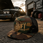 A camouflage trucker hat with a black mesh back and the text 'GET YOUR SHIFT TOGETHER' printed on the front, placed on asphalt in front of a classic car and an auto shop at sunset.