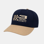 A baseball cap with a dark navy blue crown and a tan visor, featuring an embroidered logo on the front that reads '3 PEDAL SOCIETY' alongside a checkered flag design. The background is a light gray and white checkered grid pattern, indicating a digital cutout.